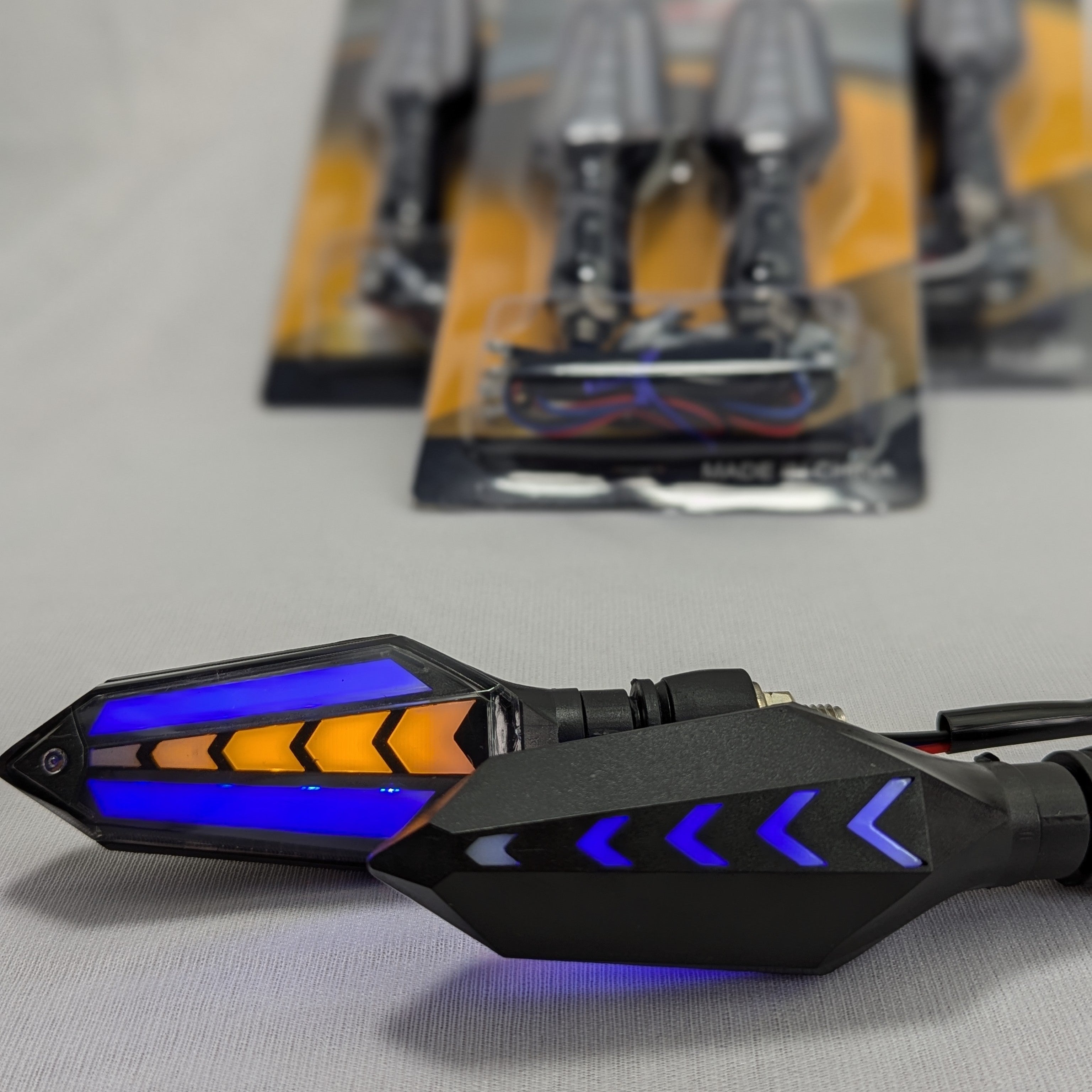 Motorcycle turn signals with blue and orange lights on a gray surface, packaging in the background.